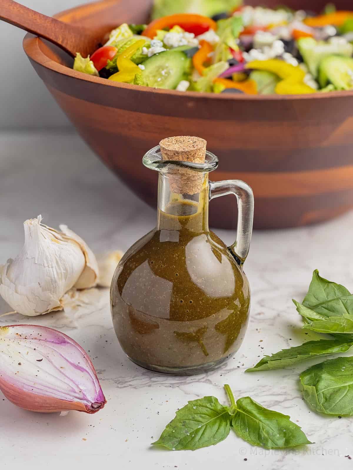 Vinaigrette in a glass carafe with a cork lid surrounded by fresh basil, shallot, and garlic cloves.