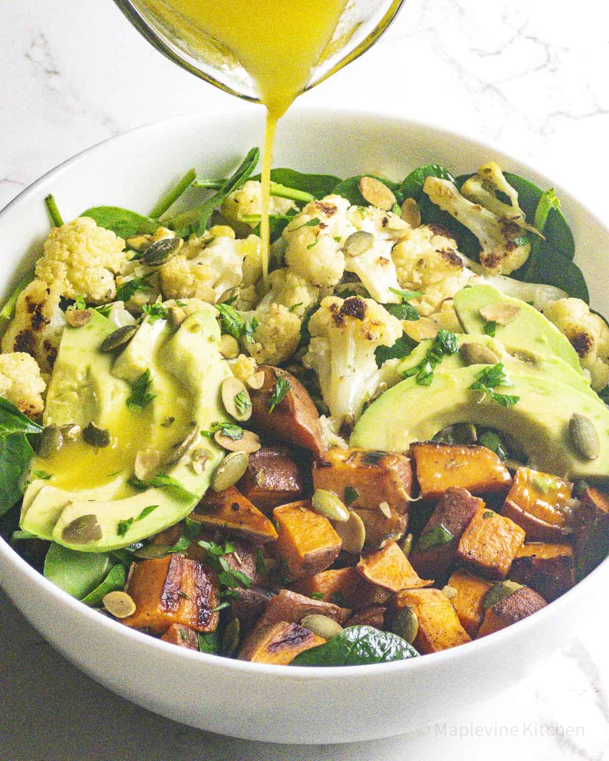 Roasted veggies, avocado slices, spinach and pepitas in a bowl with lemon dressing being poured over.