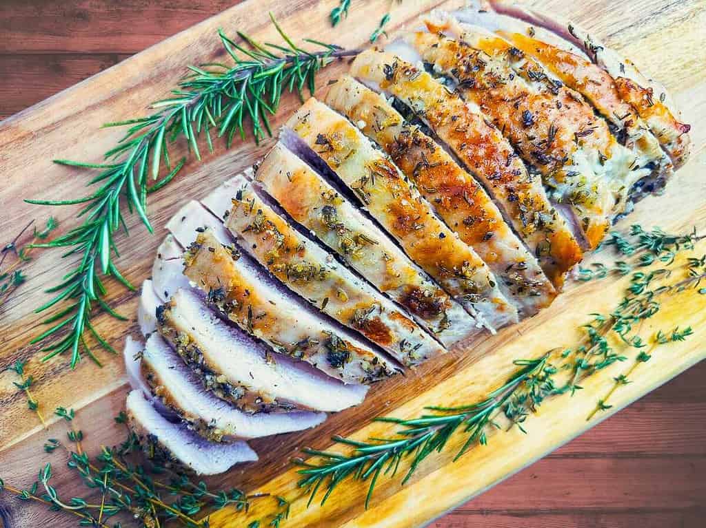 Sliced roasted turkey breast on a wooden board with fresh rosemary and thyme.