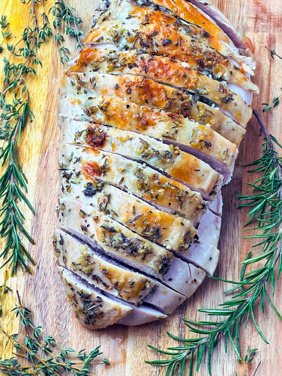Roasted turkey breast sliced on a board surrounded by fresh herbs.