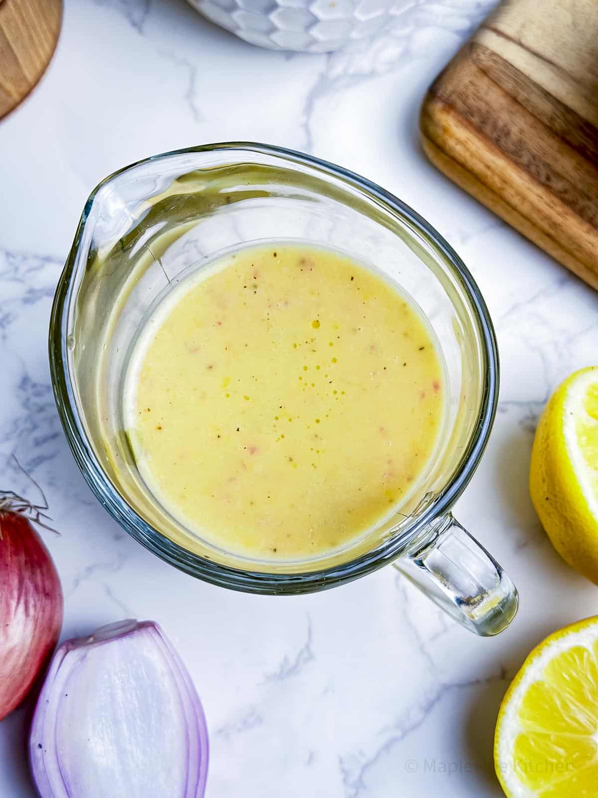 Creamy lemon dressing in a glass carafe with lemons and shallots surrounding.