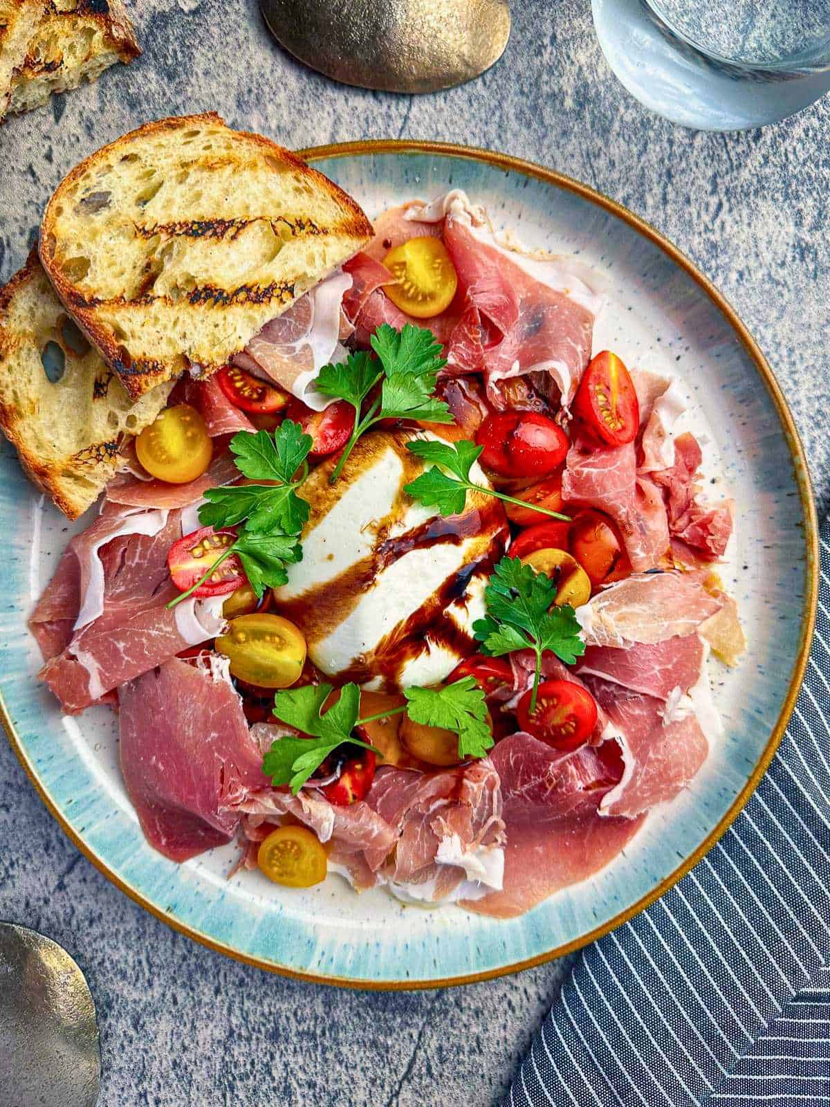 Burrata and prosciutto with tomatoes and fresh herbs and balsamic glaze on a stylish plate.