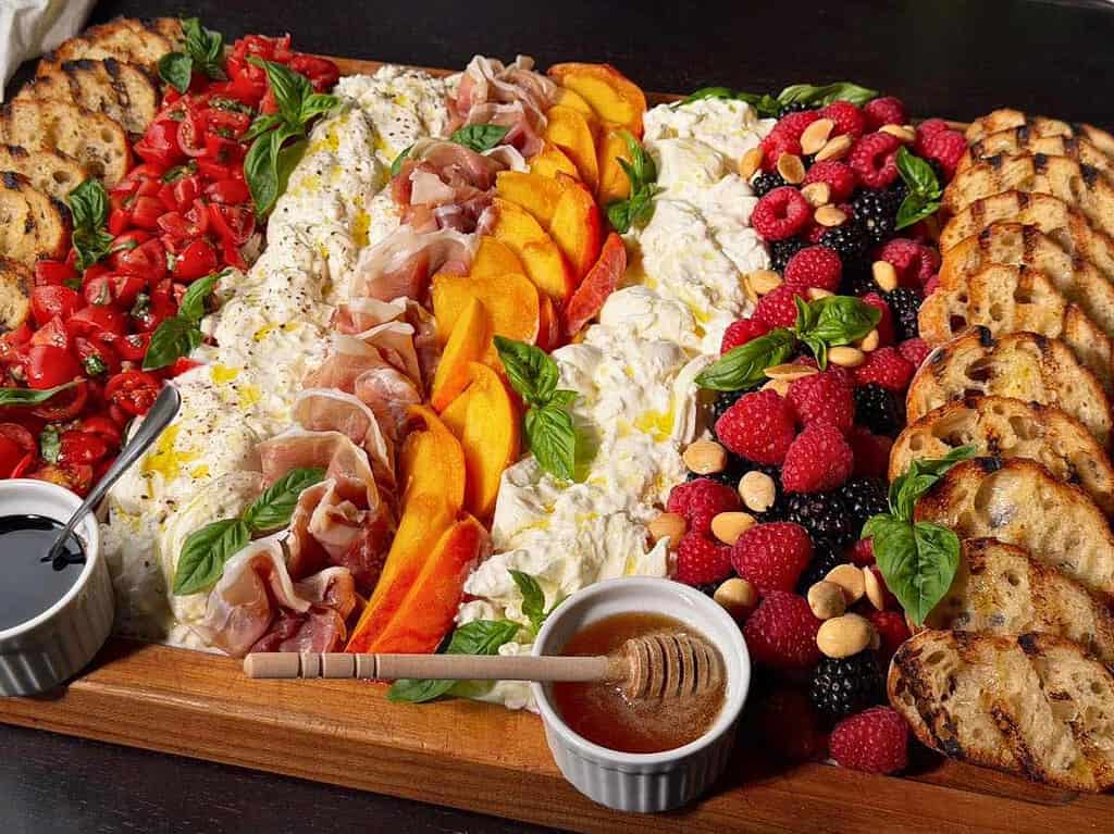 Summer Burrata Board featuring bruschetta tomatoes, peaches, prosciutto, berries, basil, and grilled crostini.