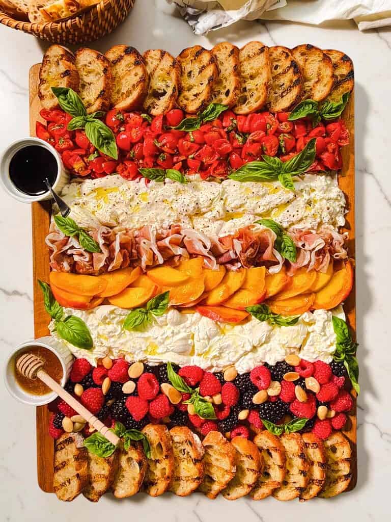 Summer Burrata Board featuring bruschetta tomatoes, peaches, prosciutto, berries, basil, and grilled crostini.