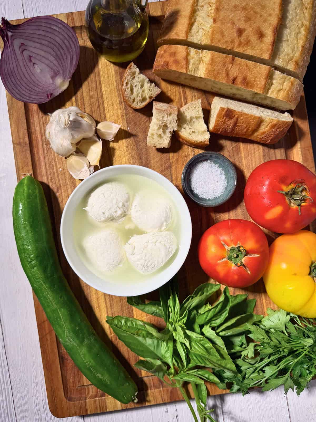 Ciabatta, tomatoes, cucumber, basil, garlic, onion, and fresh mozzarella on a wooden cutting board.