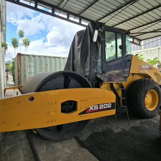 Vista de maquinaria de construcción, compactadora XS 202, en un almacenamiento cubierto, lista para trabajos de pavimentación o construcción pesada. Equipamiento robusto y eficiente para obras de gran escala.