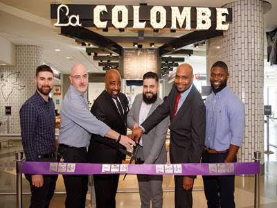 Group of six diverse men at ribbon-cutting ceremony in front of La Colombe sign.