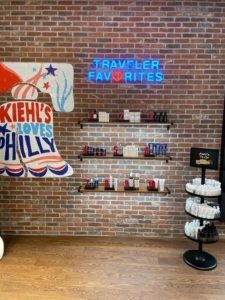 Neon sign reading 'Travel, Fave, Frites' above shelves with shoes on a brick wall.