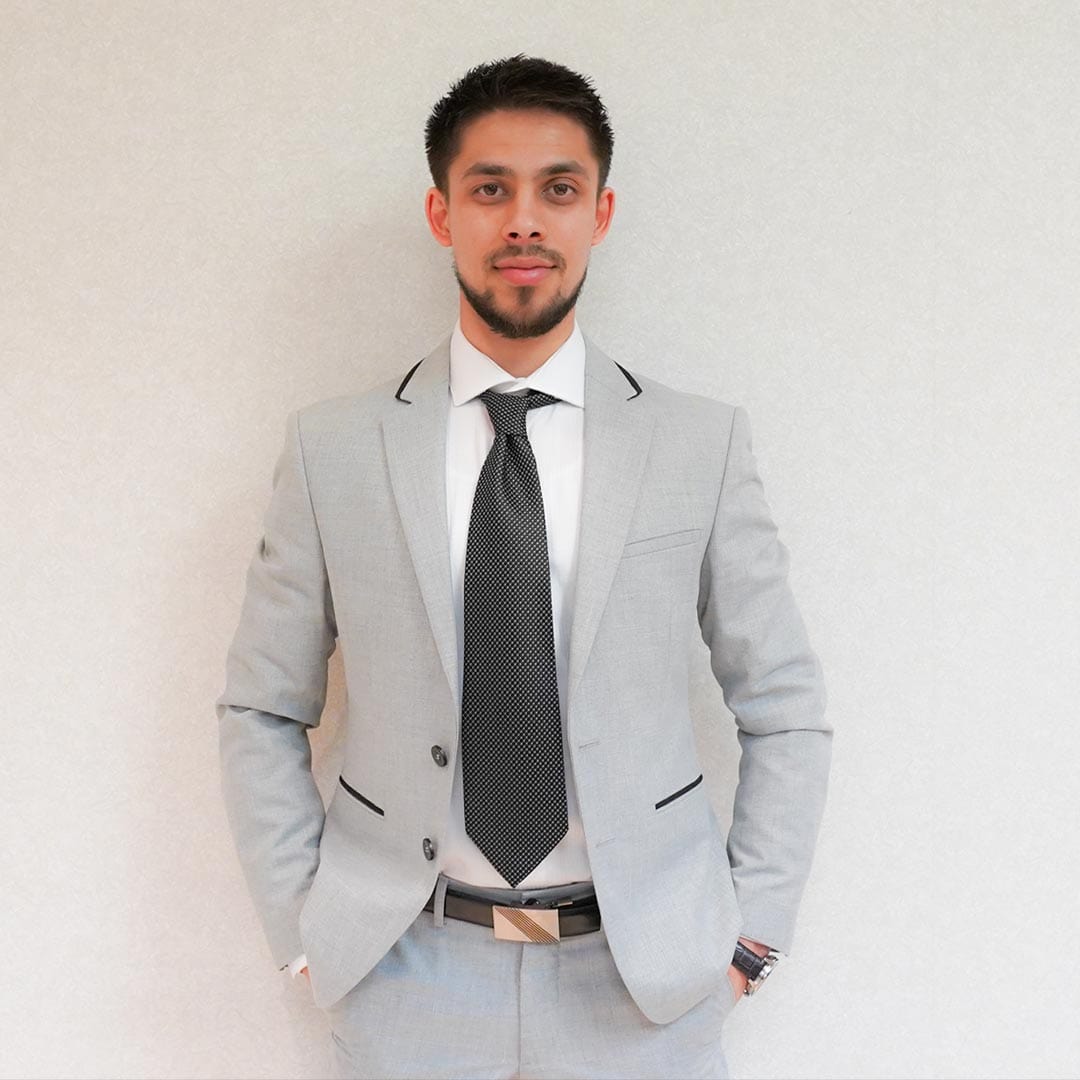 Professional man in light gray suit and black tie standing confidently against a plain wall.