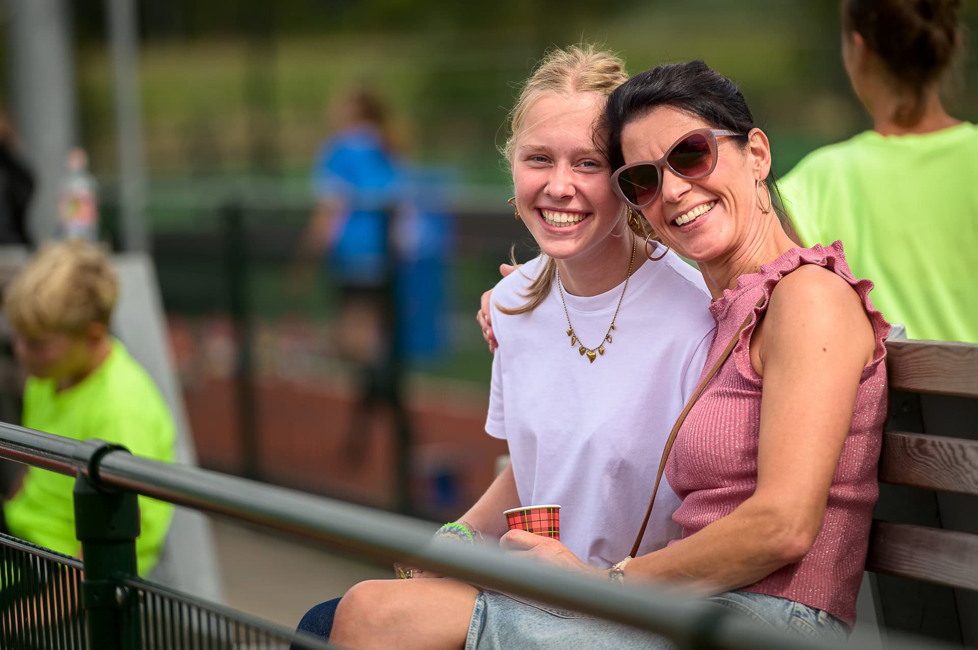 Twee vrouwen zitten op een bankje, lachend, met een hockeyveld op de achtergrond tijdens de evenfotografie van het allemanstoernooi.