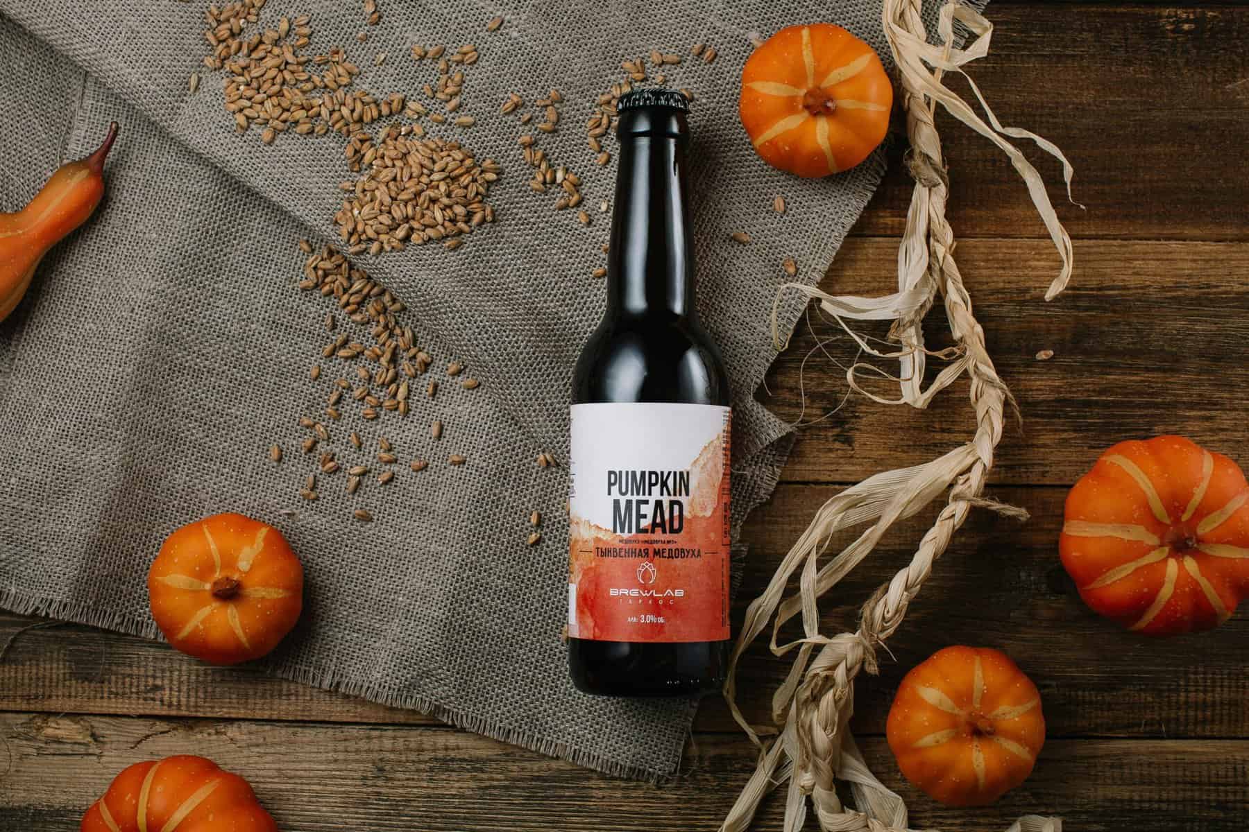 Bottle of pumpkin mead surrounded by small pumpkins and wheat grains on burlap.