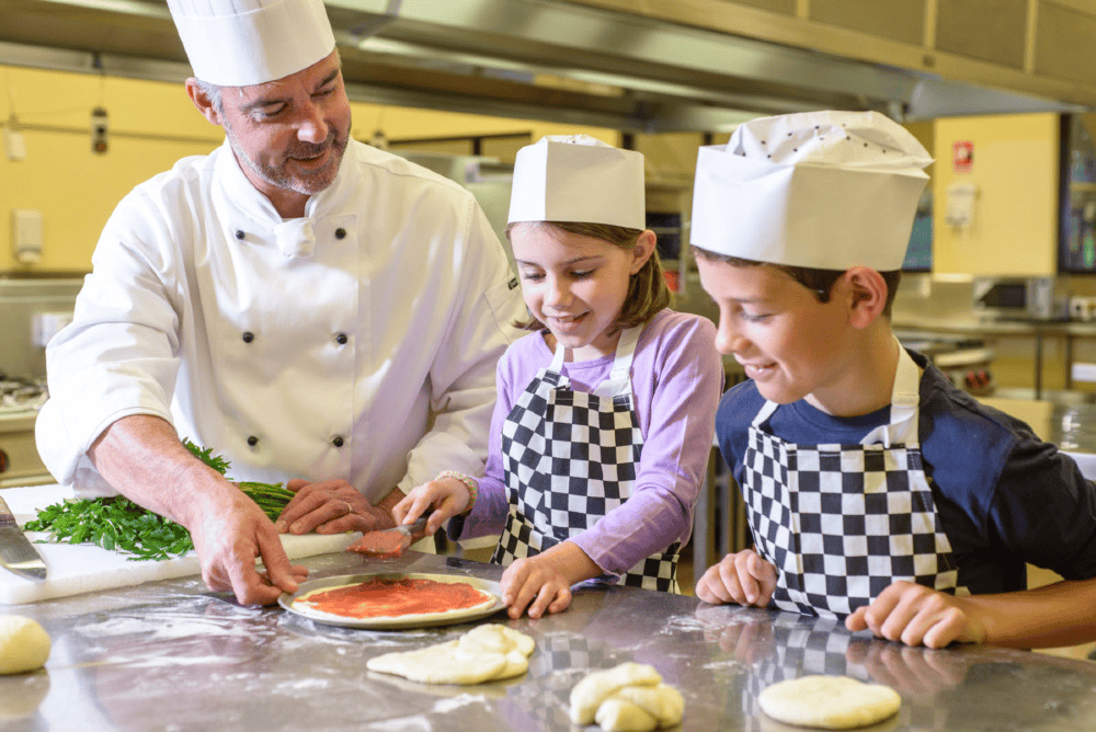 chef helping kids cook Meals in Motion Freshly made pizza with kids and chef in a commercial kitchen Kids cooking pizza with adult chef meal preparation culinary class teamwork family friendly meal service Meals in Motion