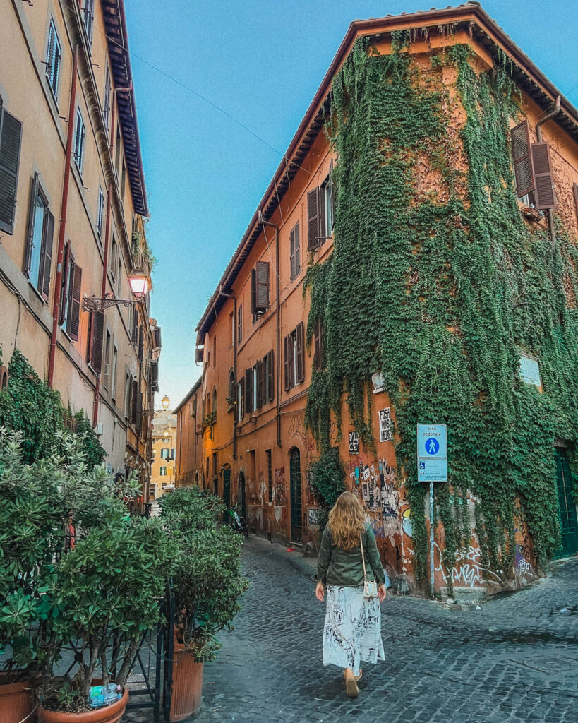 This is the beautiful neighborhood of Trastevere with this narrow street lined with cobblestone and surrounded on either side by four story buildings with a Roman color red/orange hue, brown shutters, and foliage cascading down the side. It is one of the best areas to stay in Rome for 3 days.