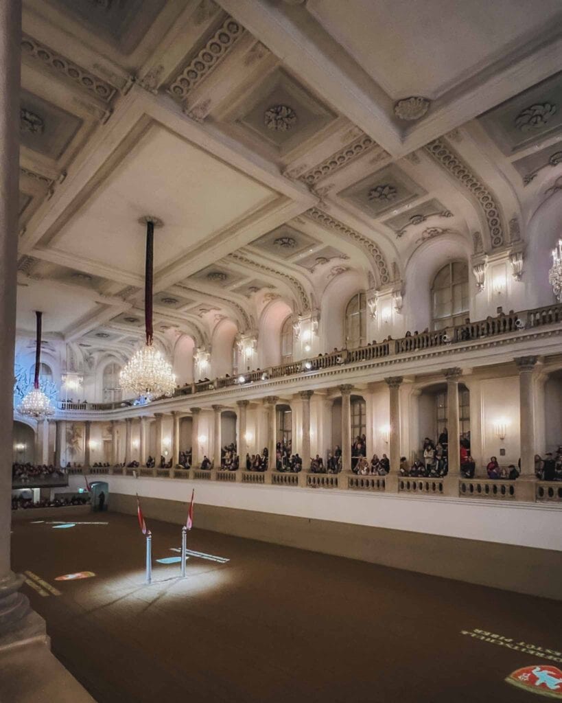 The Spanish Riding School in Vienna is famous, and you can even view one of their grand horse shows! The architecture is in the baroque style, and guests sit around the large arena in a grand, white, main hall. There are two levels to this hall with an intricate decorated ceiling, curved windows, columns, and oversized chandeliers. The horse show happens in the arena, that's actually paved with sand.