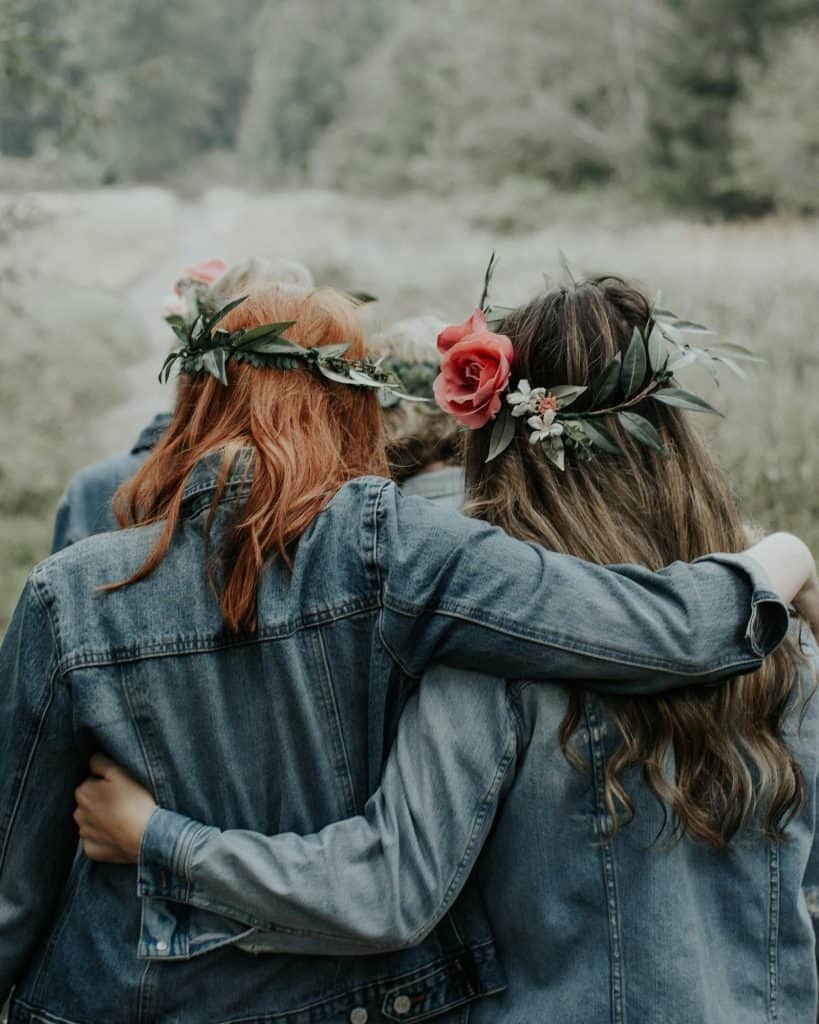 Two women walking arm in arm through a meadow, wearing flower crowns