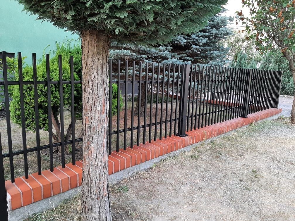 Palisadowe ogrodzenie metalowe z czarnymi prętami, osadzone na czerwonej ceglanej podmurówce, w otoczeniu drzew i zieleni - ogrodzenie Metal-Serwis Ob.