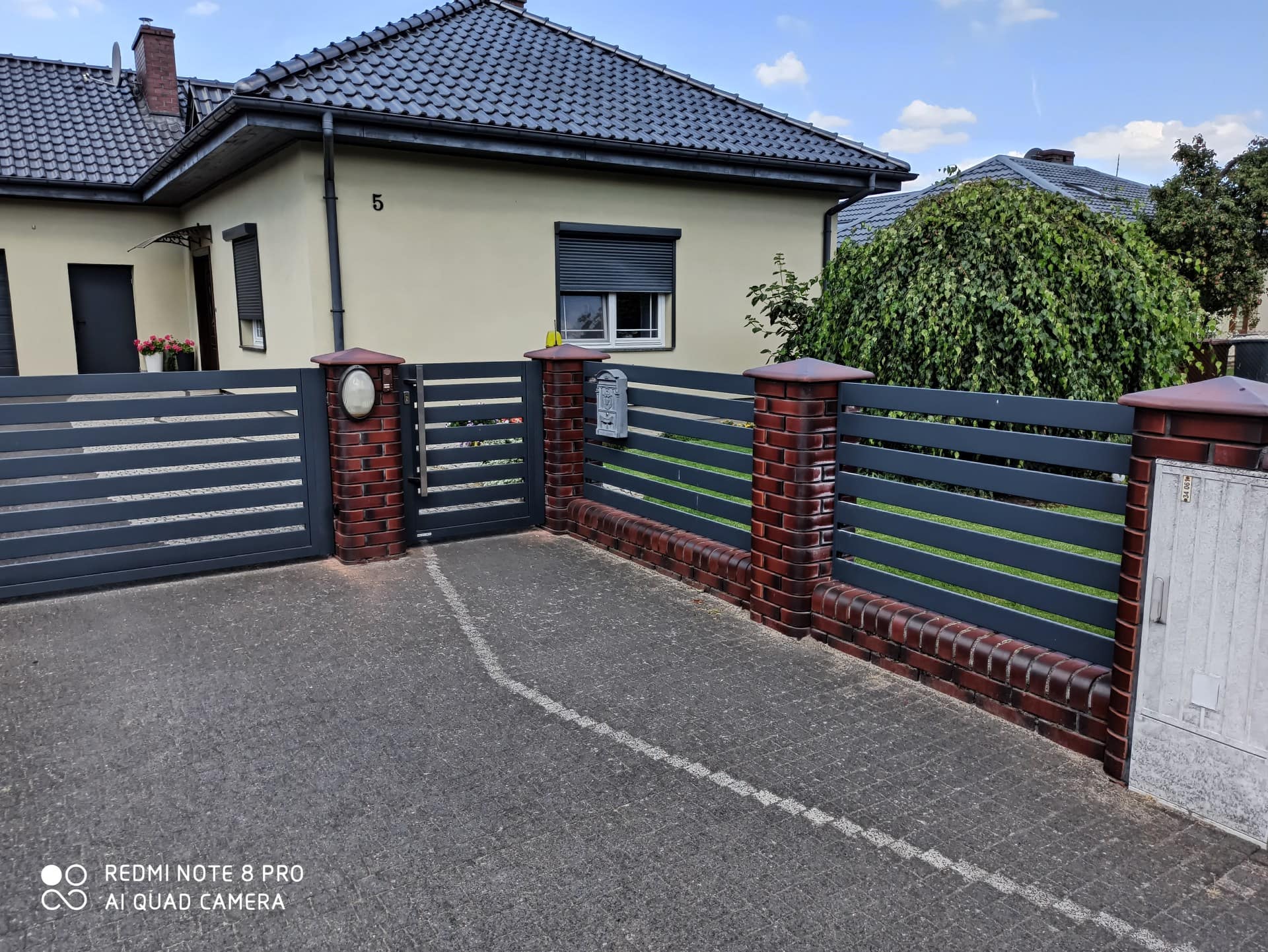 Nowoczesne ogrodzenie palisadowe z metalowych lameli, osadzone na ceglanych słupkach, w kolorze ciemnoszarym - ogrodzenie Metal-Serwis Obrzycko.