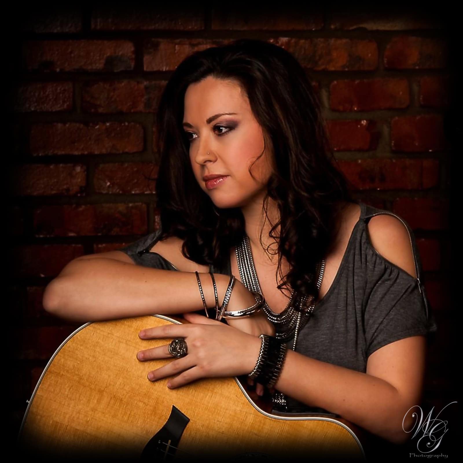 Portrait of a female guitarist with dark hair and jewelry against a brick wall.