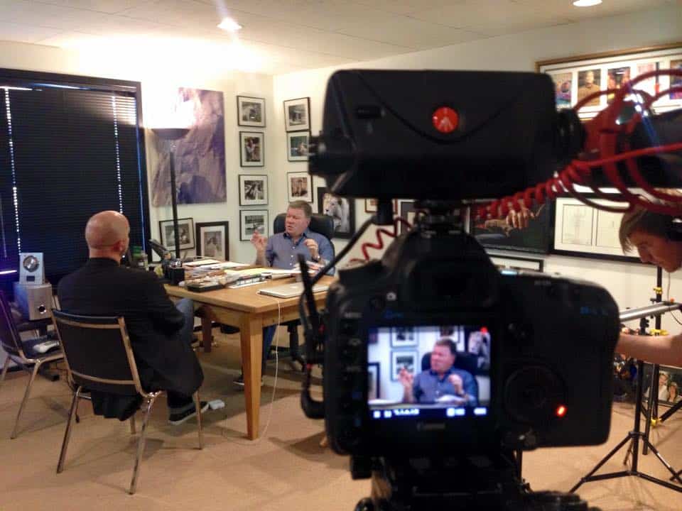 Interview scene with two men seated at a desk, camera recording the conversation.