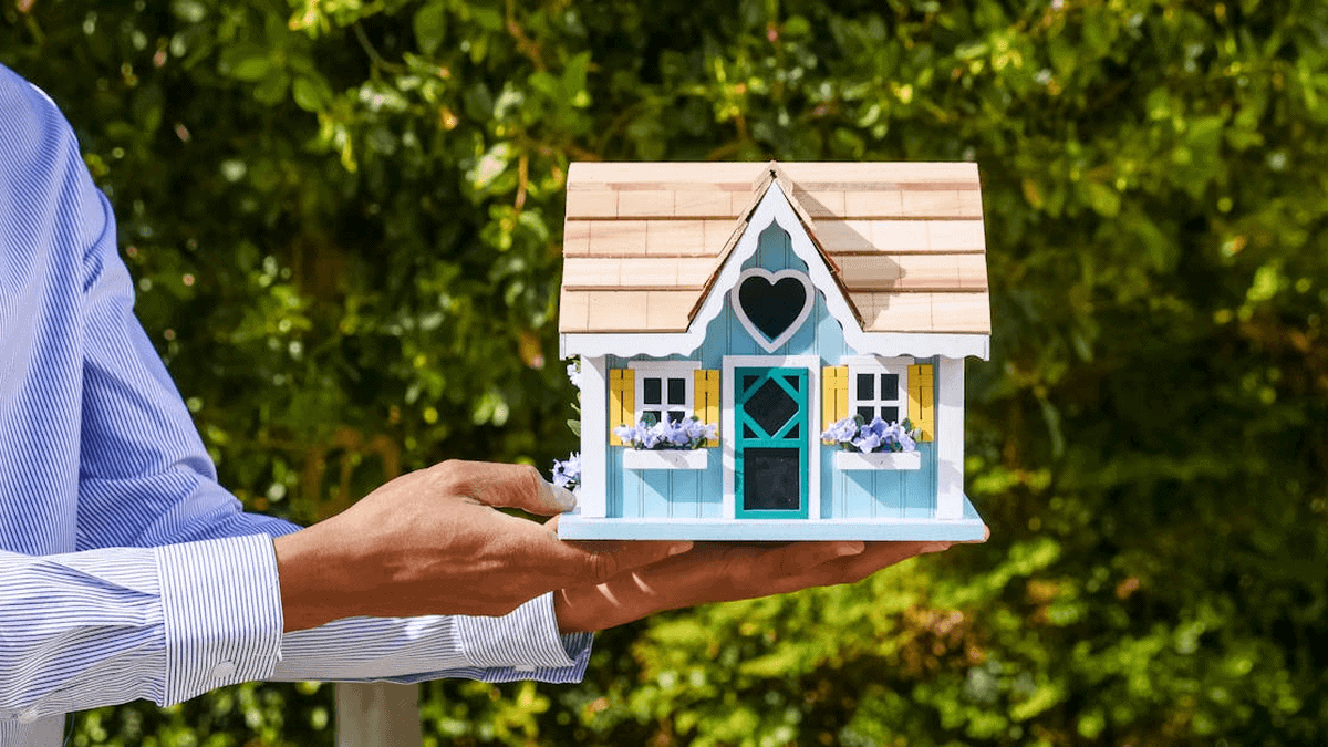 Hand holding a charming miniature house model with a heart-shaped window, set against a lush green b.