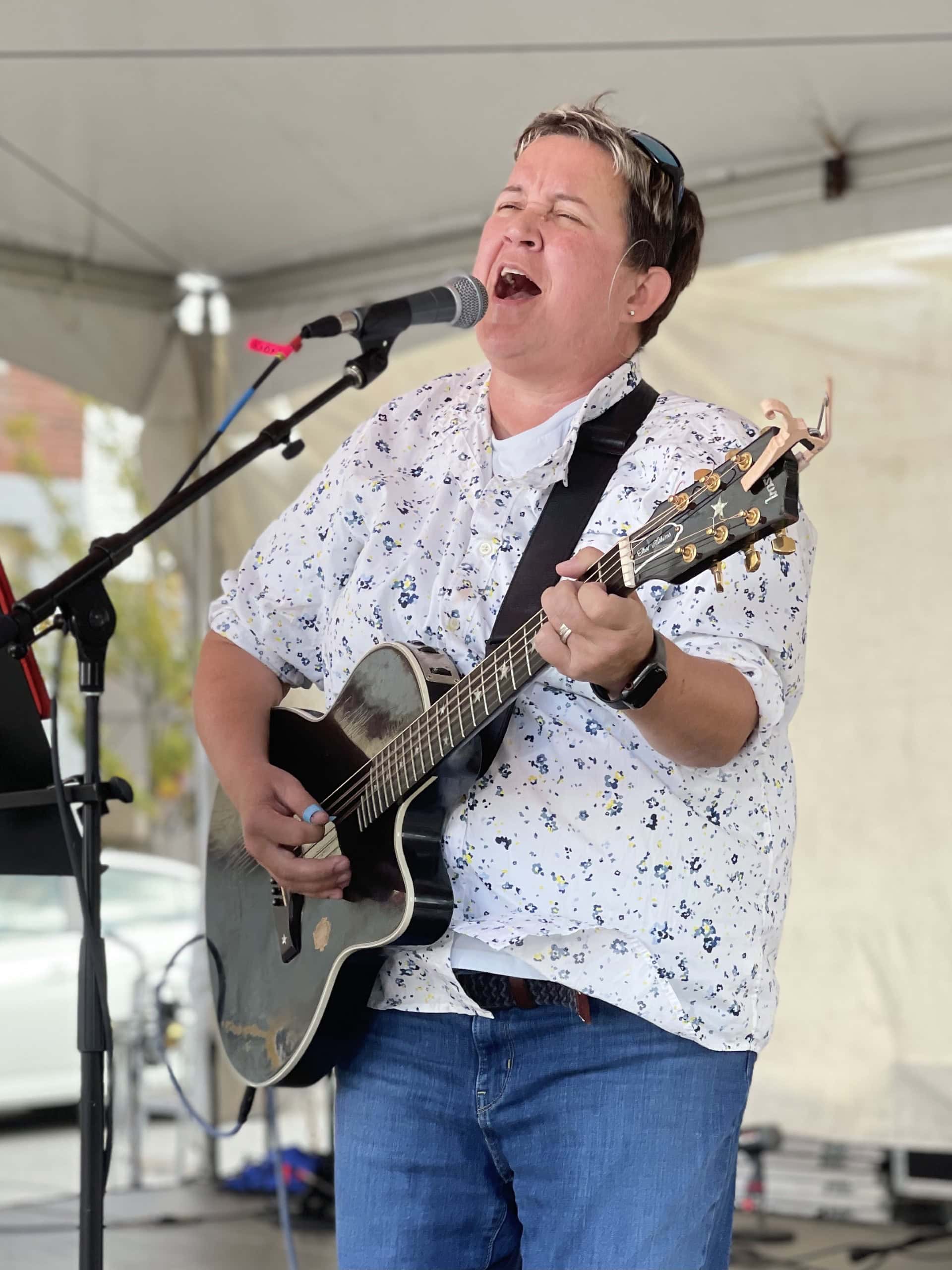 Woman singing and playing guitar at outdoor event under a tent.