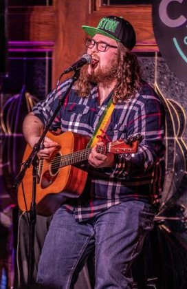 Musician with long hair and glasses playing guitar and singing into a microphone.