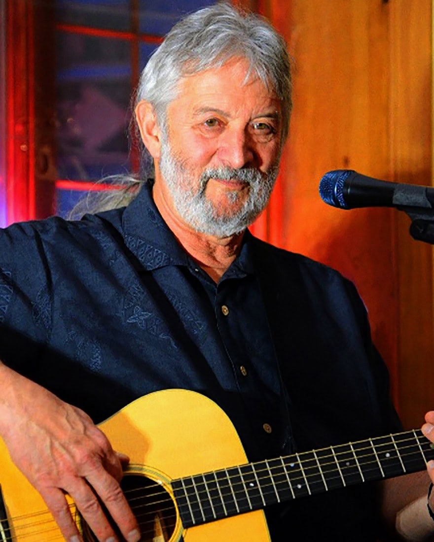 Older man with gray hair and beard playing guitar and singing into a microphone.