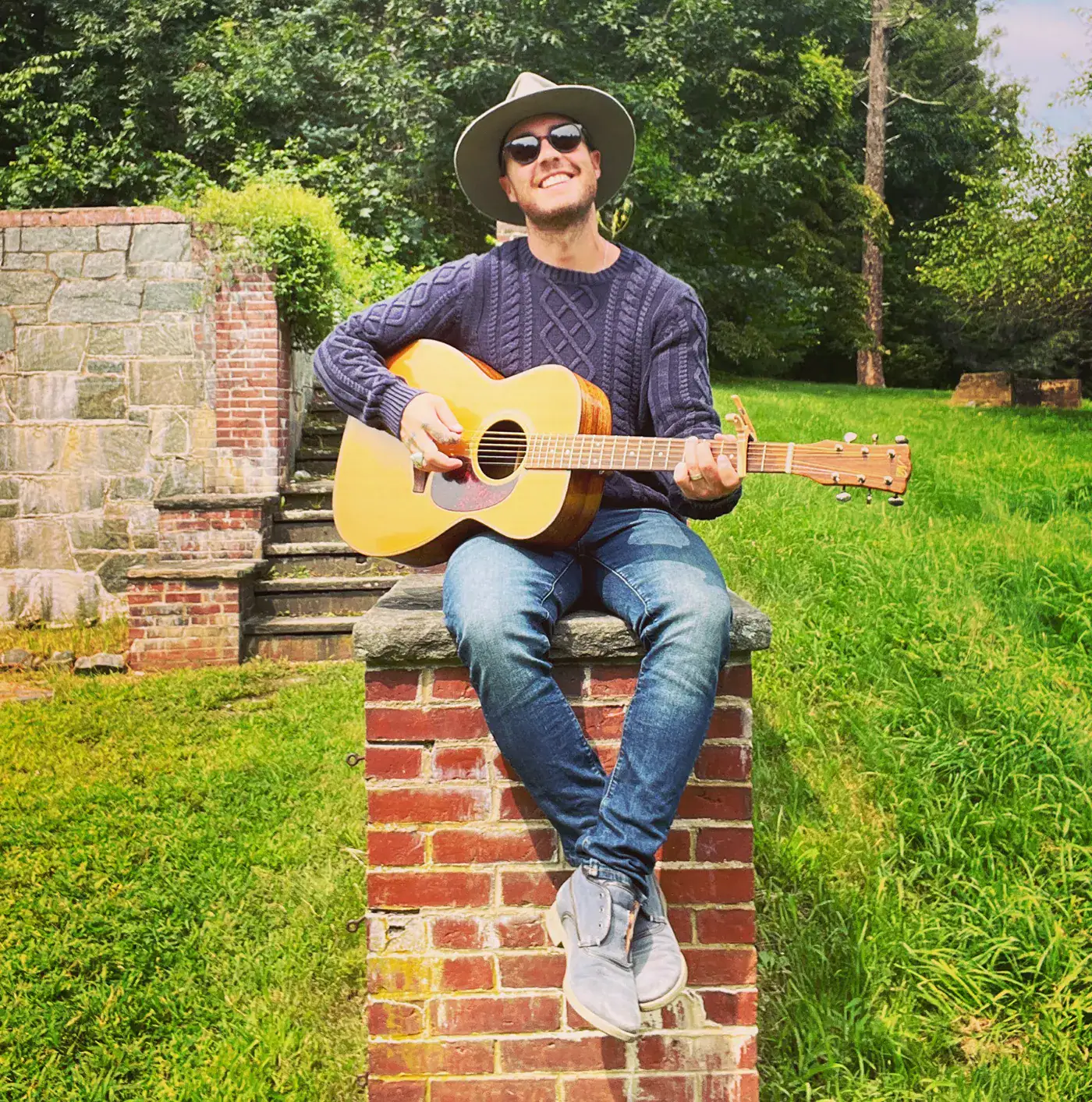 Man with sunglasses and hat playing guitar outdoors on brick structure in green park.