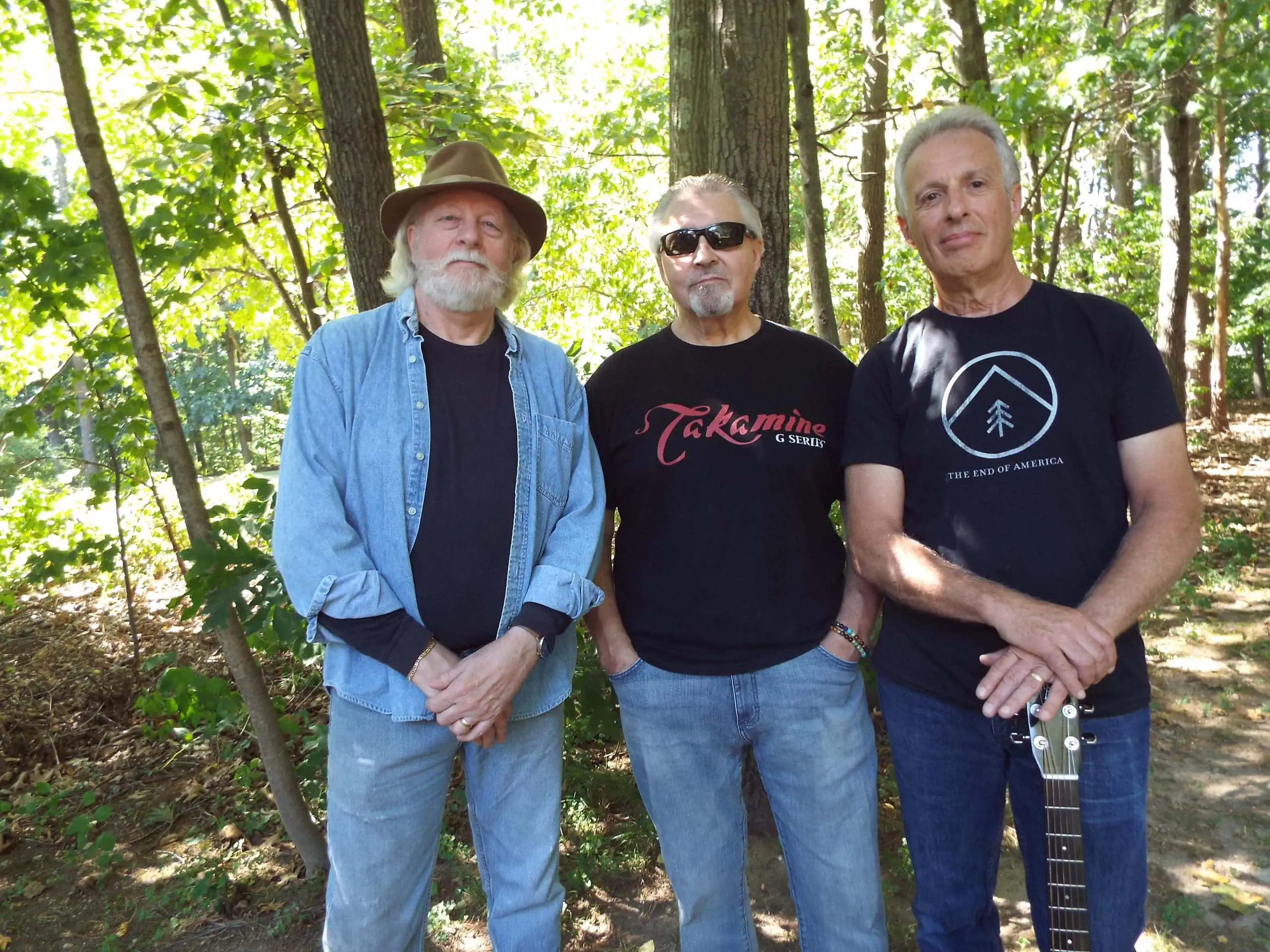 Three men standing outdoors in a wooded area with sunlight filtering through trees.