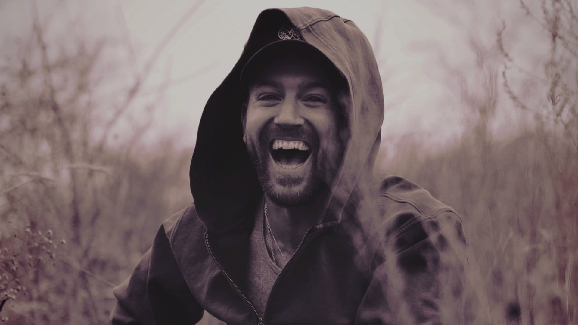 Man smiling outdoors in a hooded jacket with trees in the background.