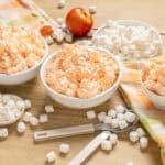 Creamy marshmallow fluff in bowls with scattered mini marshmallows, apples, and colorful checkered cloth on a wooden surface, perfect for dessert recipes or sweet treats.