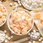 Creamy marshmallow and sweet potato casserole in white bowls on a wooden table with marshmallows scattered around and a colorful checkered cloth.