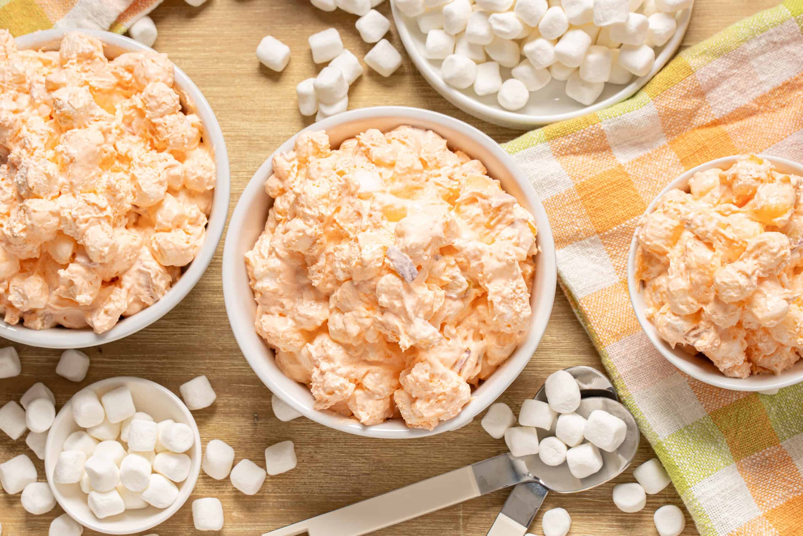 Creamy marshmallow and sweet potato casserole in white bowls on a wooden table with marshmallows scattered around and a colorful checkered cloth.