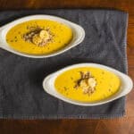 Creamy yellow soup garnished with bread crumbs and herbs served in white oval bowls on a dark cloth and wooden tabletop.