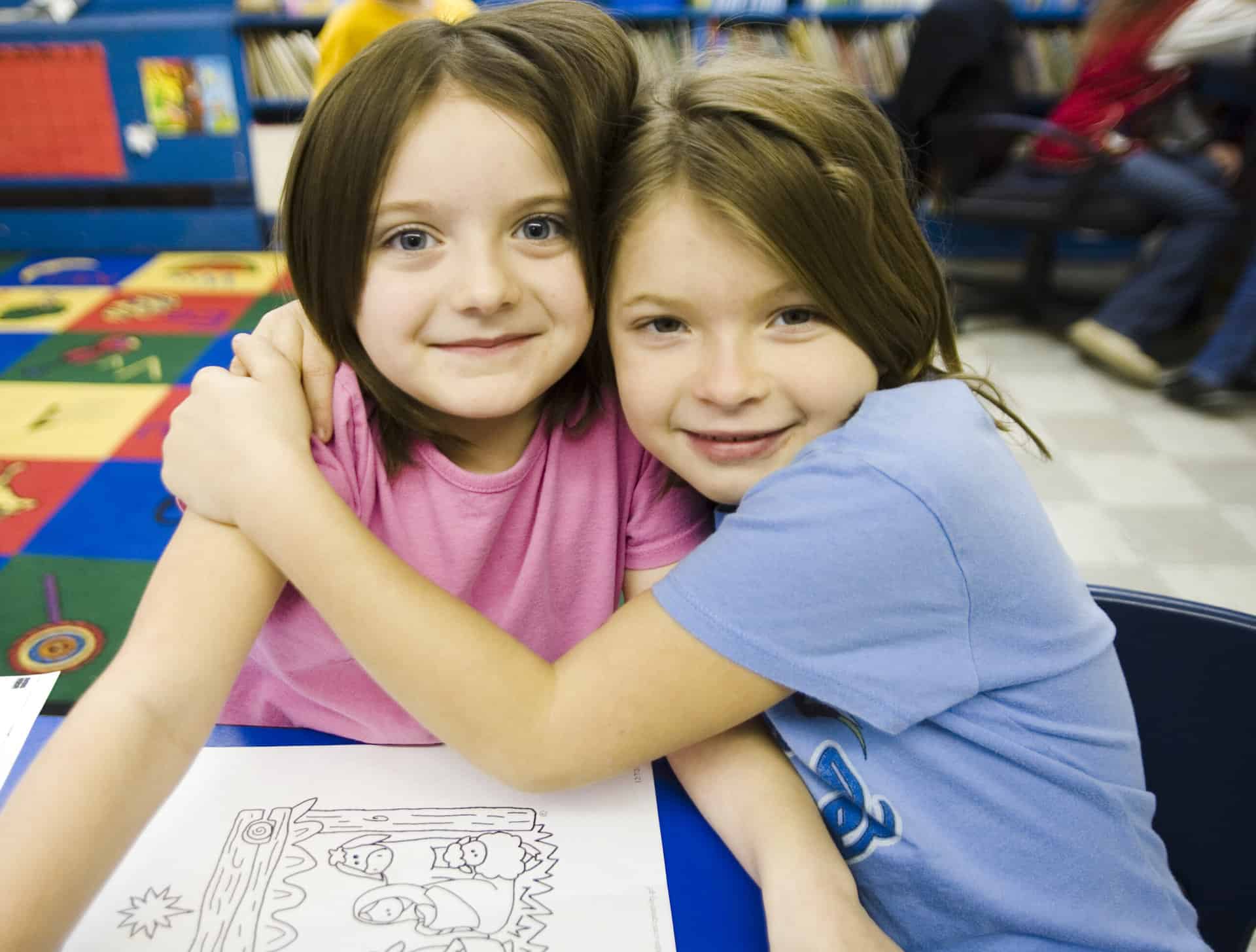 Two young girls hugging at a table in a classroom relying on back to school programs.