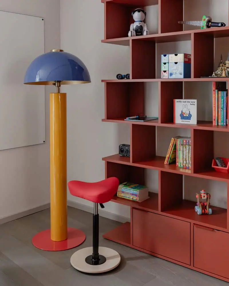 Blue and yellow modern floor lamp beside a red bookshelf filled with toys and books in a contemporar.