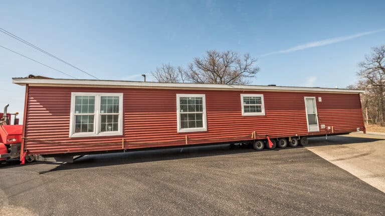 Red mobile home on a paved lot with trees in the background.