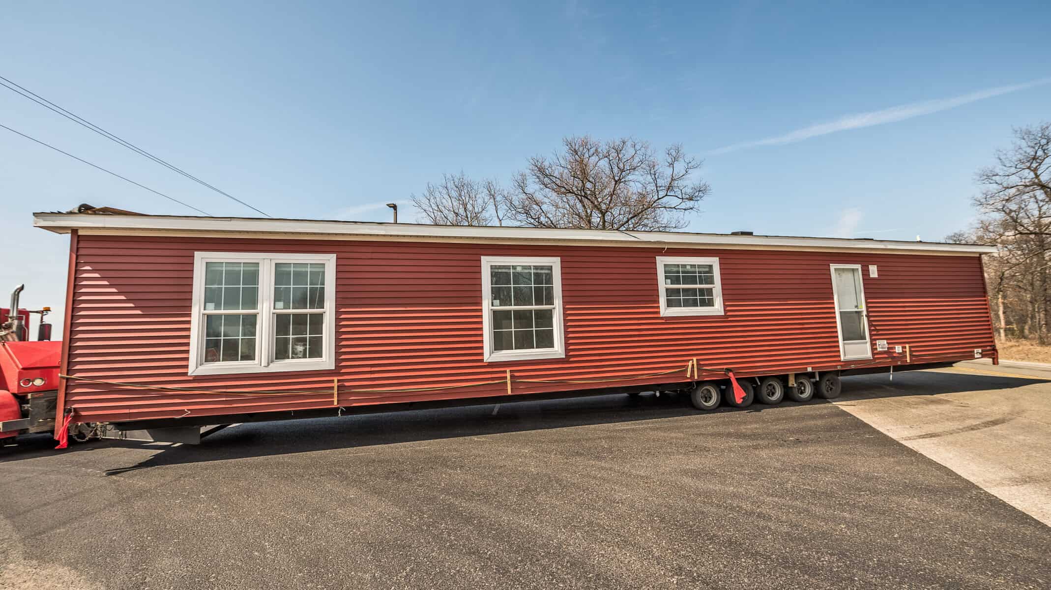 Red mobile home on a paved lot with trees in the background.