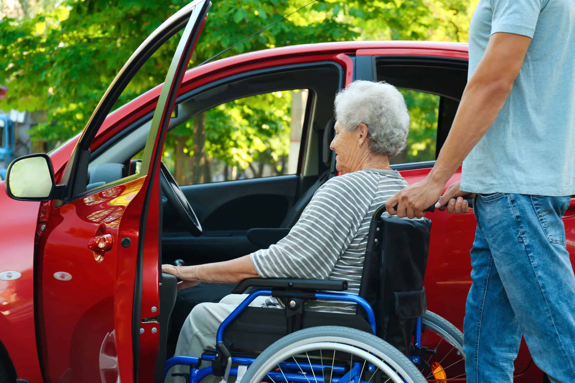 Young man helping disabled senior woman in wheelchair to get into car outdoors