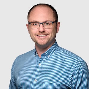 A professional headshot of a smiling veterinarian wearing glasses and a blue checkered shirt, standing against a light gray background.