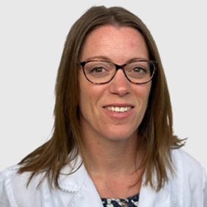 Professional headshot of a smiling veterinarian with long brown hair wearing glasses and a white lab coat over a patterned blouse, against a plain, light background.
