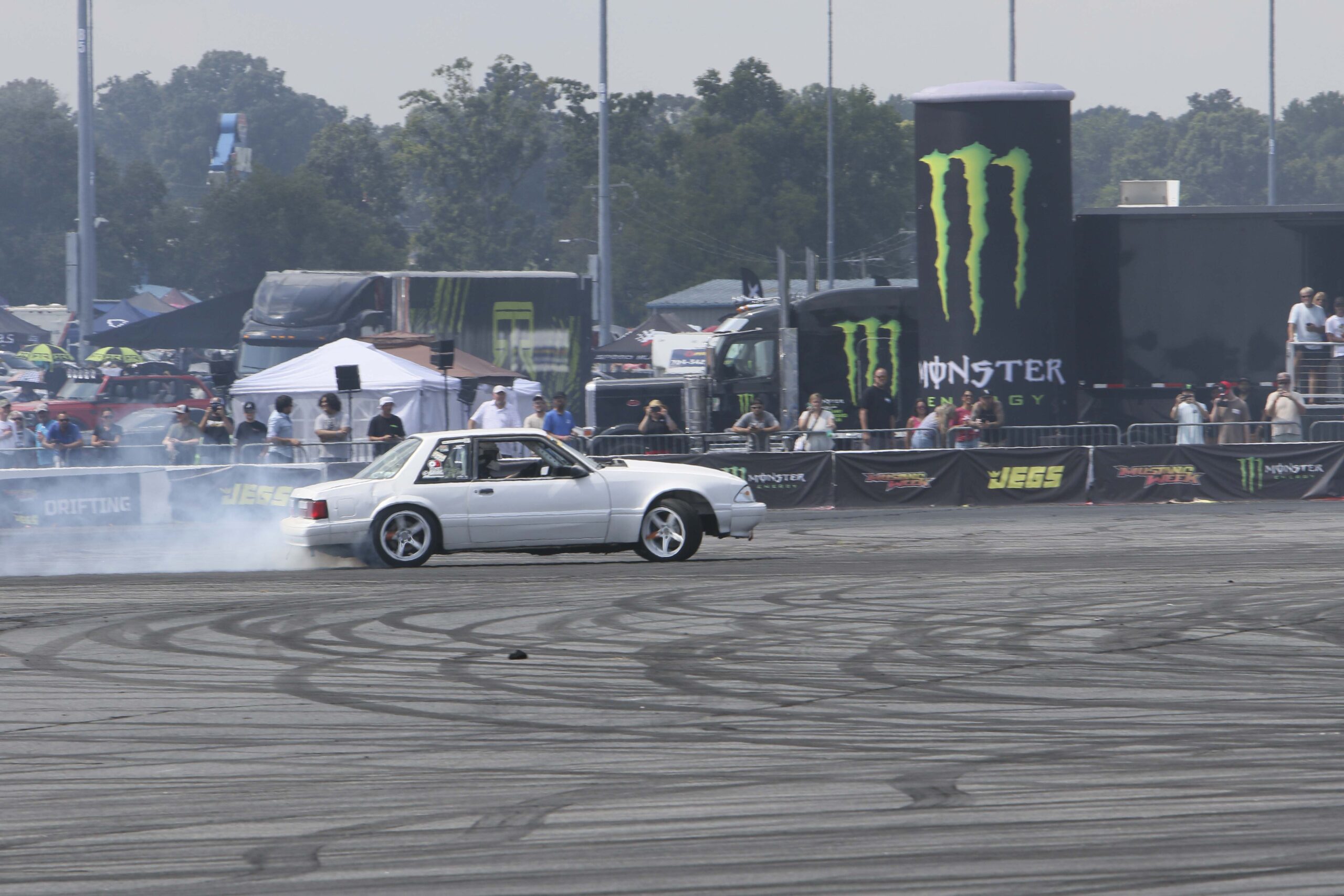 White Mustang drifting at Mustang Week event with tire smoke and crowd in background.