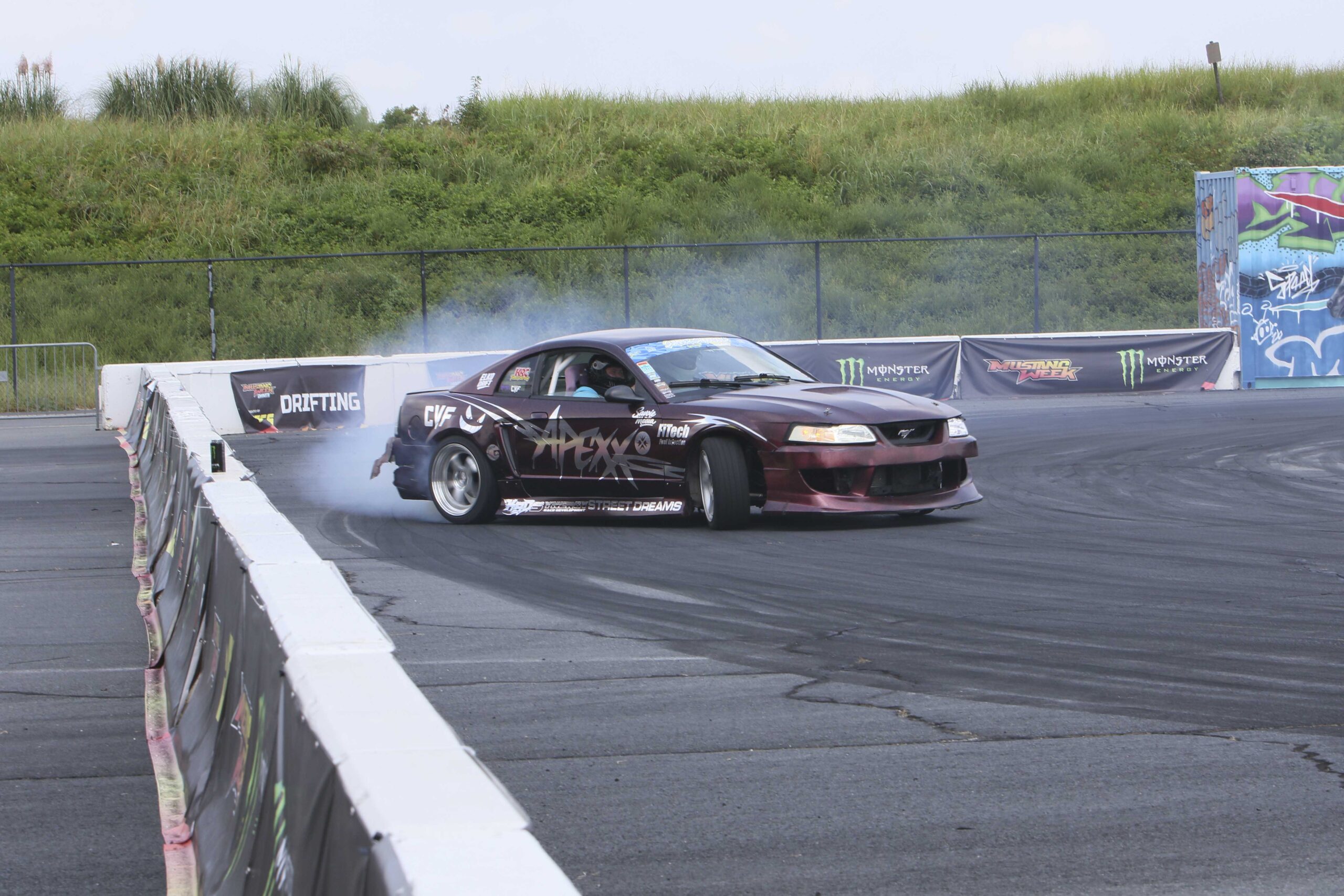 Drifting Mustang at Mustang Week with tire smoke and track barriers.