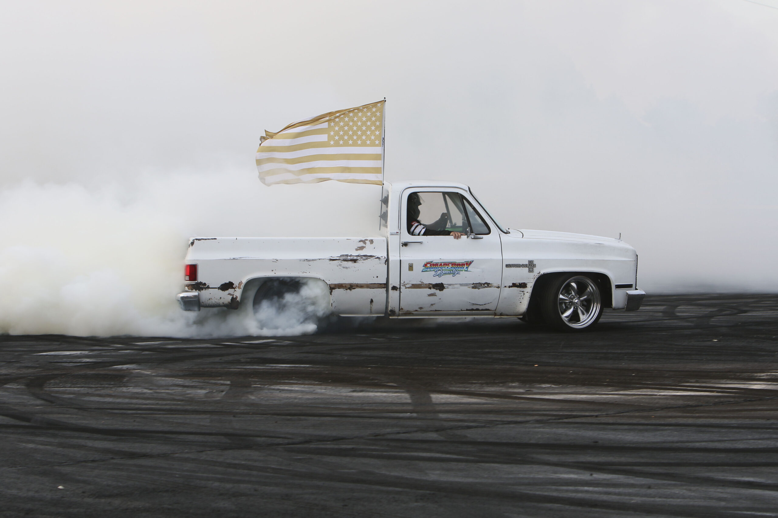 White pickup truck performing a burnout with American flag at Mustang Week event.