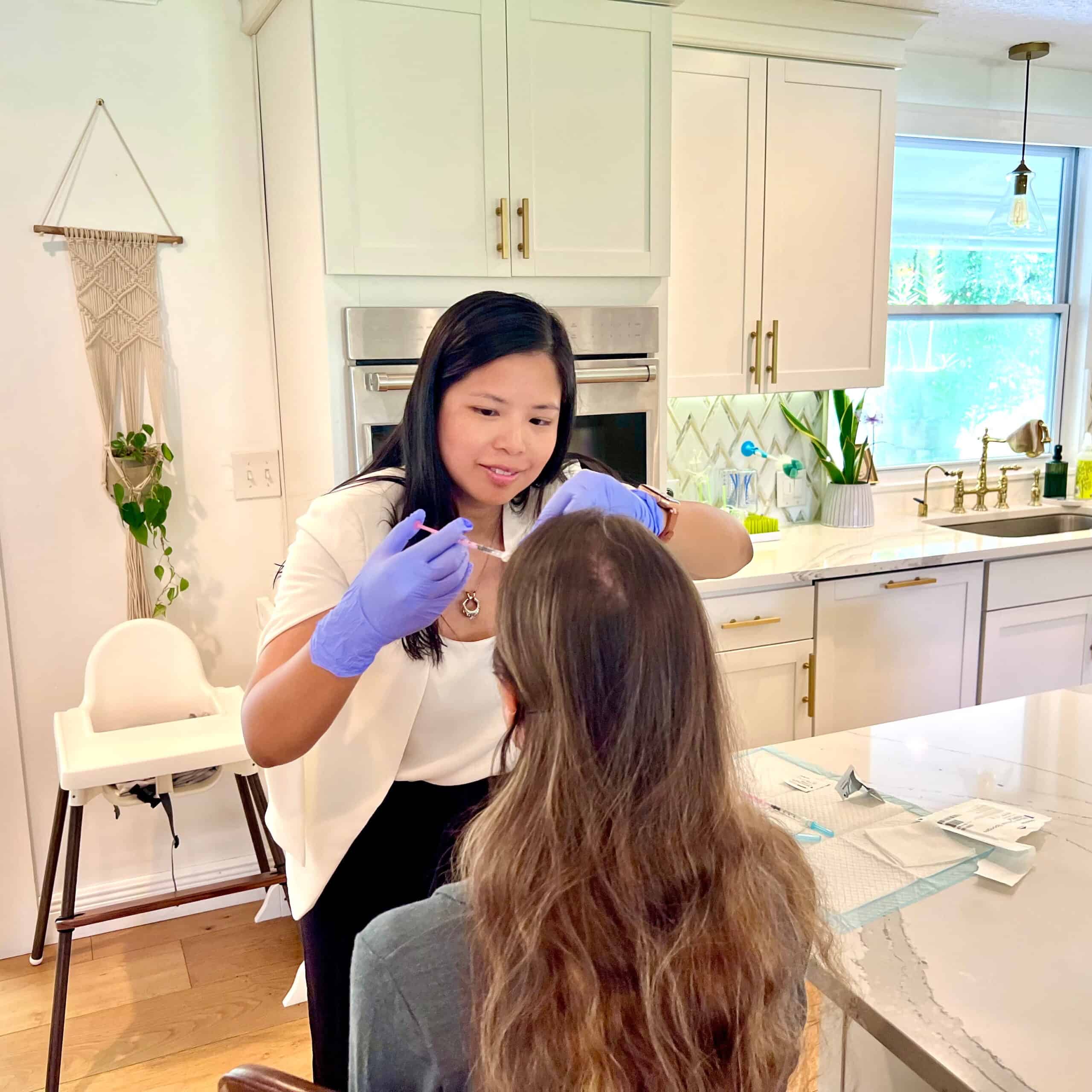 Dr. Samantha Mikals injecting wrinkle relaxer to the forehead of a brunette long haired woman sitting at her kitchen counter.