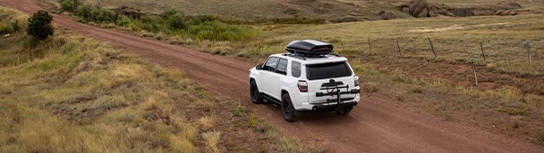 White SUV with roof cargo carrier on rural dirt road in open countryside.