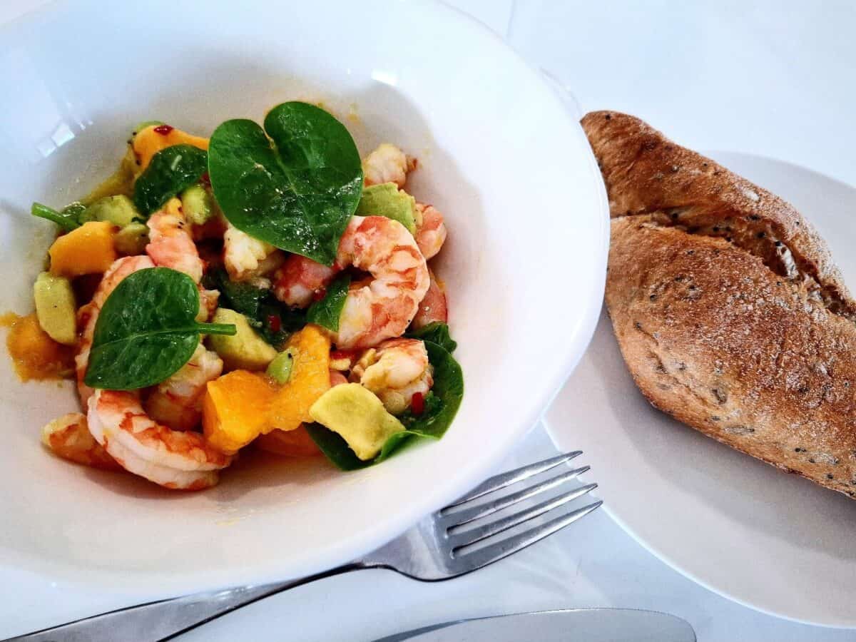 Salad prawn, baby spinach, mango and avocado in a white bowl plate on a white table. A white side plate with a wholegrain bread roll next to it and a silver knife and fork