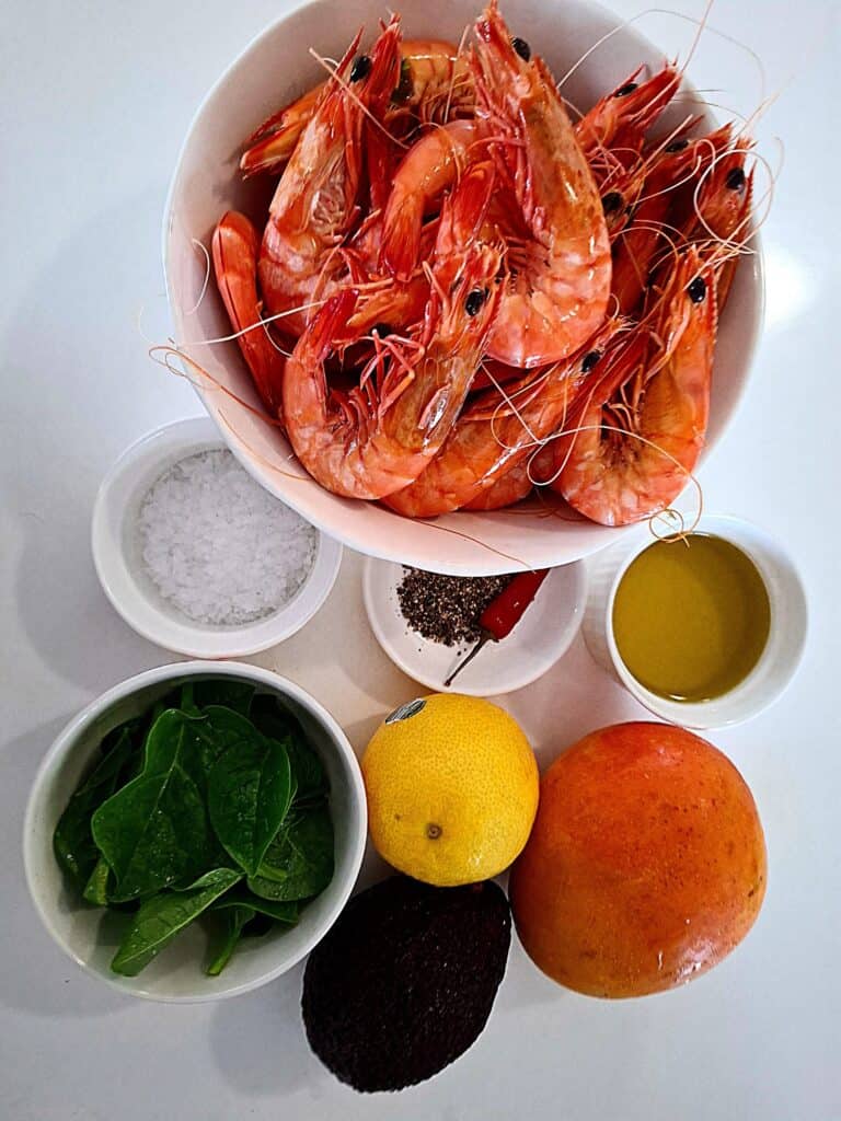 A white bowl of whole prawns in their shell on a white table, 4 small white ramekins next to it, one with spinach leaves, another with olive oil, another with cracked pepper and a chilli, and another one with flaky salt. Next to it is a whole mango, avocado and lemon