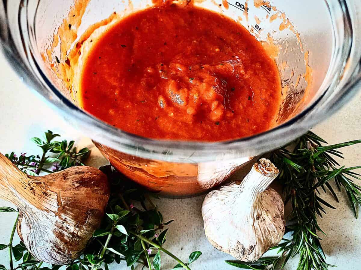 Pizza sauce in a clear blender base. Two heads of garlic and fresh herbs in front of it