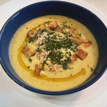 Roasted garlic cream soup topped with croutons, grated Parmesan cheese, chopped parsley and a drizzle of truffle oil. In a blue bowl on a white plate